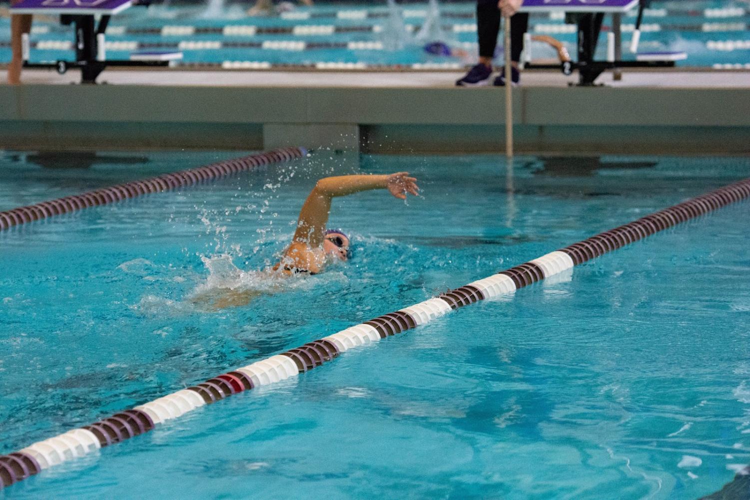 A diver wearing goggles swims away toward the end of the pool, with one arm out of the water. 