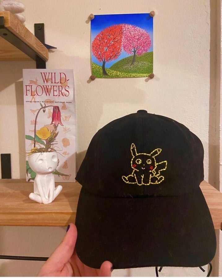 Kubis holds a black hat in her hand with a yellow Pikachu embroidered on it. She holds the hat in front of her white wall that has a poster with two orange and pink trees.