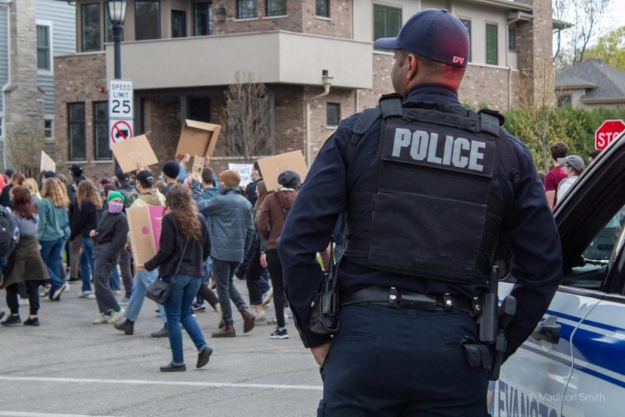 An EPD officer in a navy blue uniform, seen from behind, leans against a cop car and watches a group of protestors in the background.
