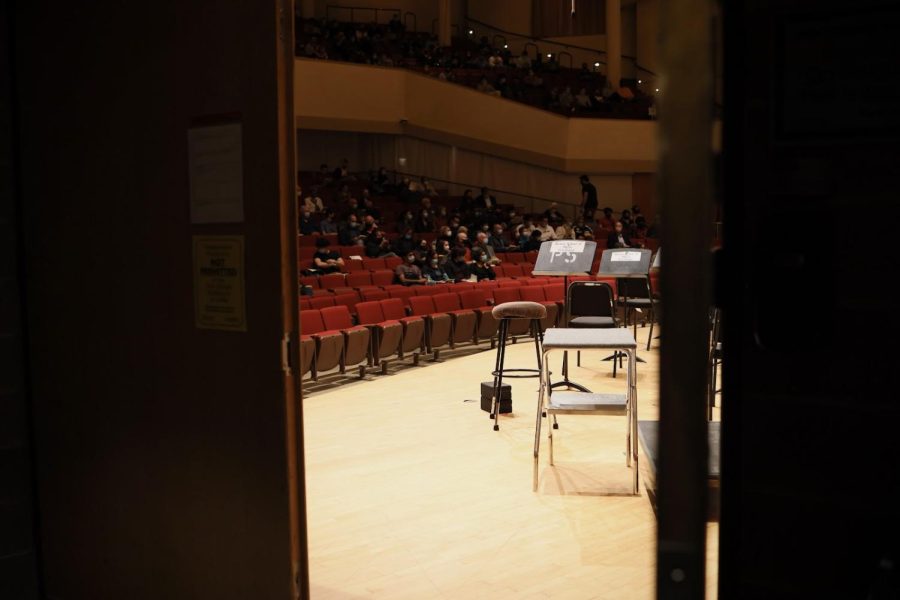 A stool sits in front of a blank music stand on an empty stage. People are visible in the red audience seats behind.