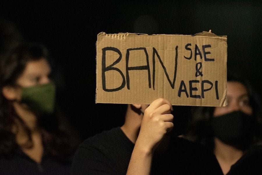 A hand holding up a sign reading BAN SAE & AEPI with faces out of focus in the background.