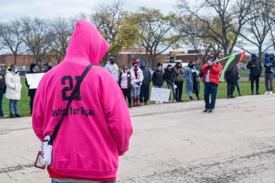 A Bost family member wears a pink hoodie with the name “Ryan Bost” and “21.” Behind them is a line of people, some holding signs, and a person with a flag and a bullhorn.
