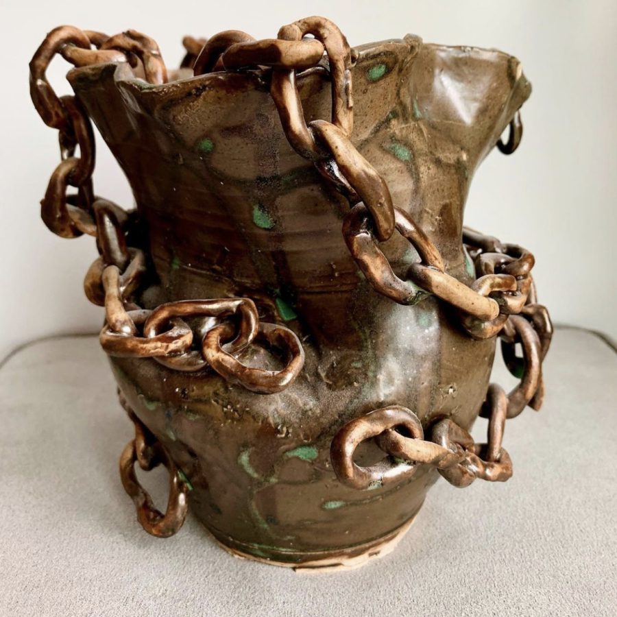 A brown vase with chains on it sits on a table. The vase has green spots.