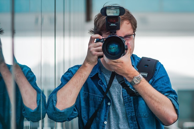 A man with short brown hair in a blue button down shirt with the sleeves rolled up holds up a camera with a large flash. He is wearing a watch on his left arm, and a bag is strapped over his shoulder. On the left, there is glass where his arm is reflected.
