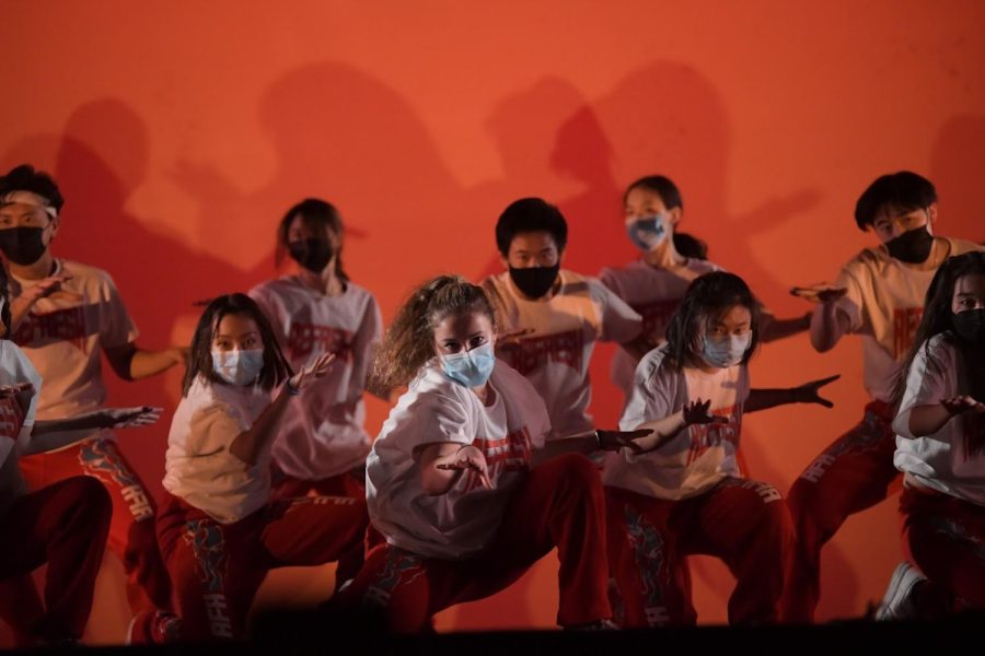 A group of multiple dancers crouch down while wearing matching white shirts with red text and red sweatpants on an orange backdrop while wearing masks.