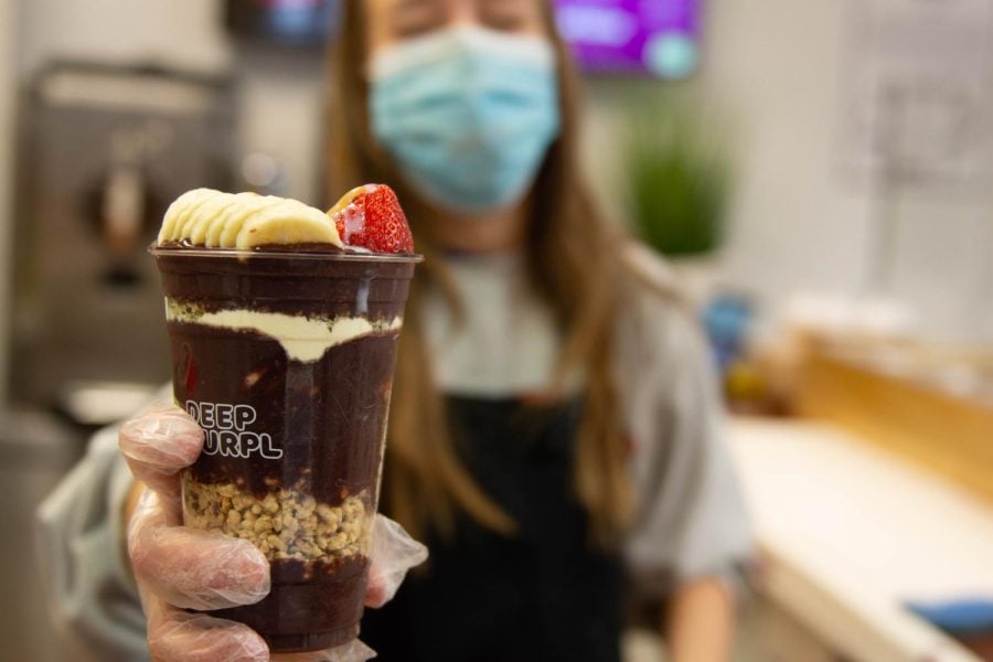 An employee with long hair wearing a blue disposable face mask holds up a clear plastic cup filled with açaí mixture and layers of granola, topped with bananas and strawberries.