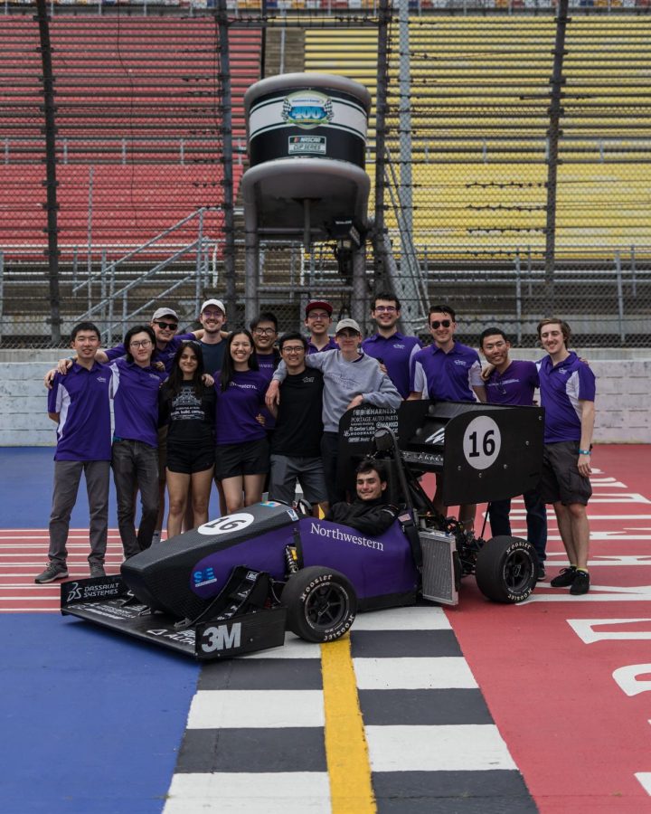 Formula poses with last year’s race car. One student sits in the purple vehicle, and a group of students stand behind the car.
