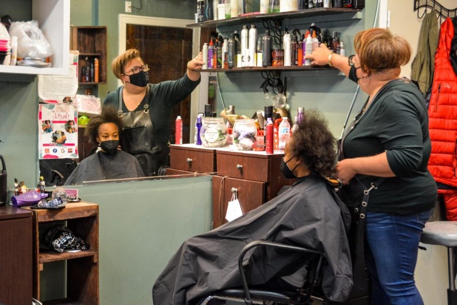 A person in a black shirt combs the hair of a client wearing a black robe. Both people are in front of a hair station with a variety of hair products.
