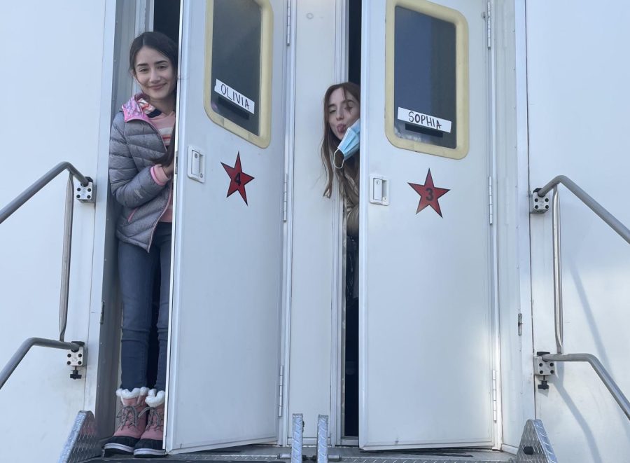 Two teenage girls, one wearing a grey jacket, jeans, and pink snow boots, and one with a blue disposable face mask, stand in the doorways of a white trailer. The doors are decorated with a red star and display the names “Olivia” and “Sophia.”