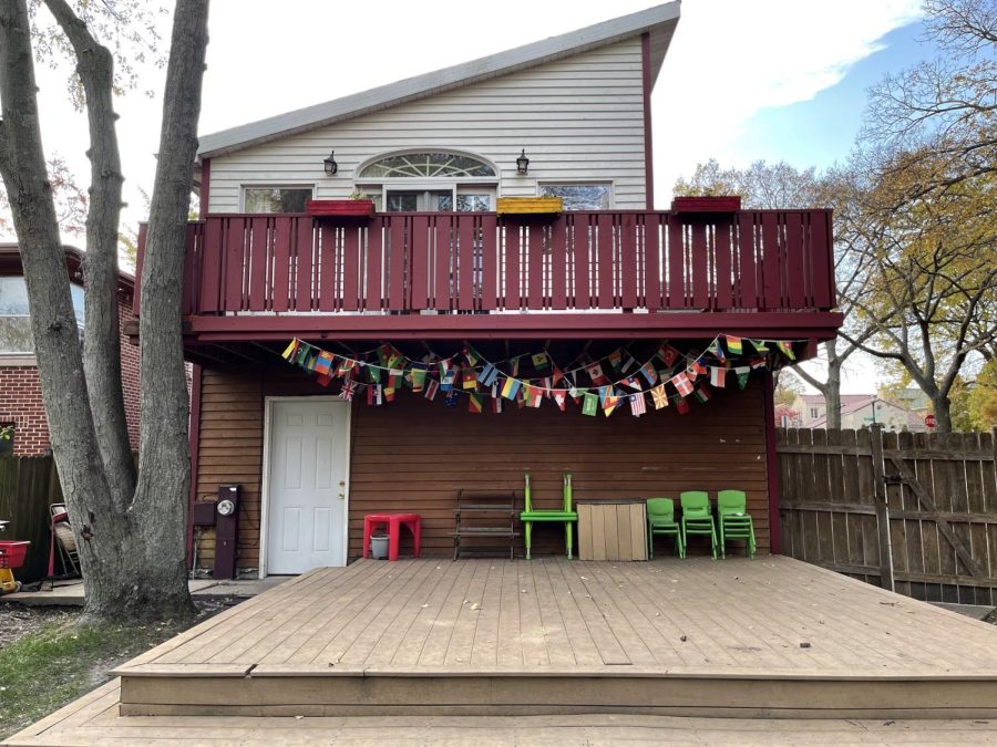 The image features the back entrance of the Evanston Montessori Children’s House. The school appears to be two levels, with a red balcony on the second floor. Below the balcony are well over 50 different flags representing the various countries of the world. Small green chairs for children are also below the balcony.