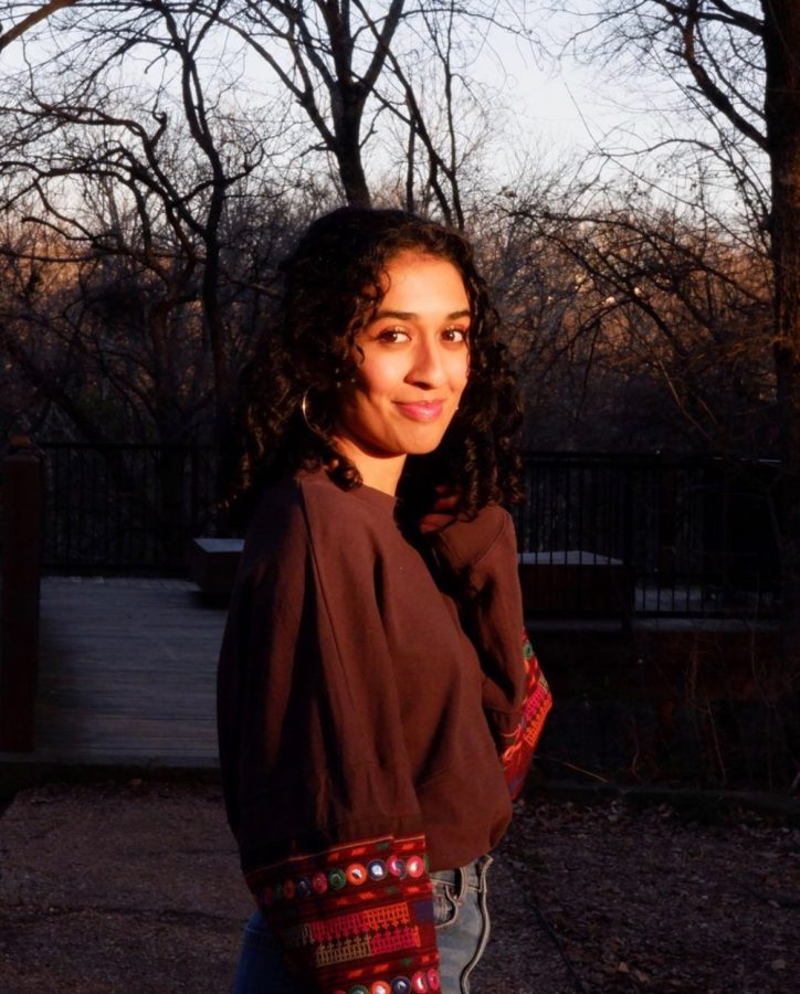 A girl in a maroon sweater with curly black hair looks at the camera with trees in the background.