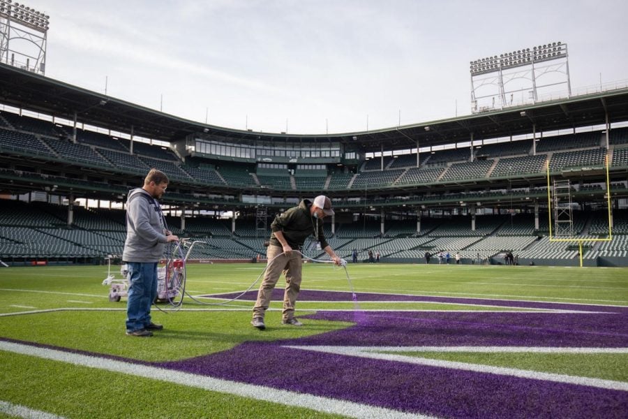 An individual holds a paint sprayer, spraying purple paint onto the center of the football field. Another individual holds the cord attached to the sprayer.