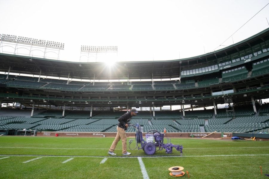 An individual pushes a purple machine on wheels on the field, with green seating behind.
