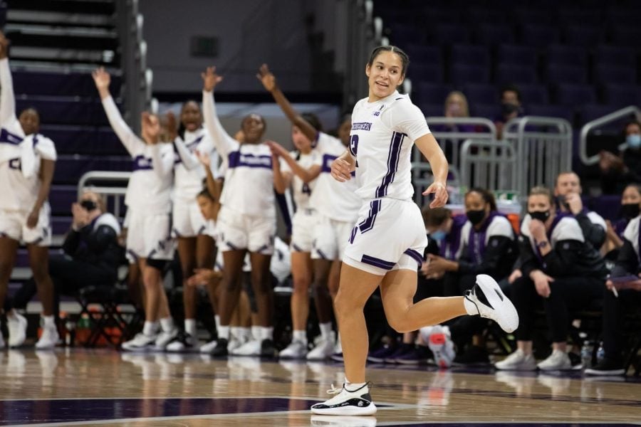 A girl in a white jersey with a purple number “12” on the front and white shorts with a purple stripe down the side looks backward on a basketball court. Multiple girls are standing with their arms up in the background.