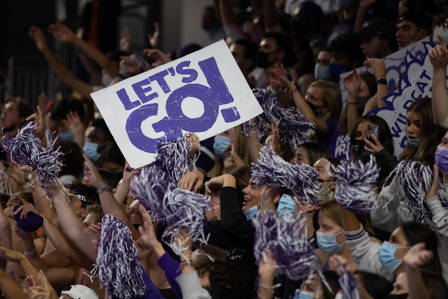 Large+group+of+students+with+arms+in+the+air+holding+purple-and-white+poms+cheer+on+the+team.+One+student+is+holding+a+white+sign+with+large+purple+letters+saying+%E2%80%9CLet%E2%80%99s+go%21%E2%80%9D