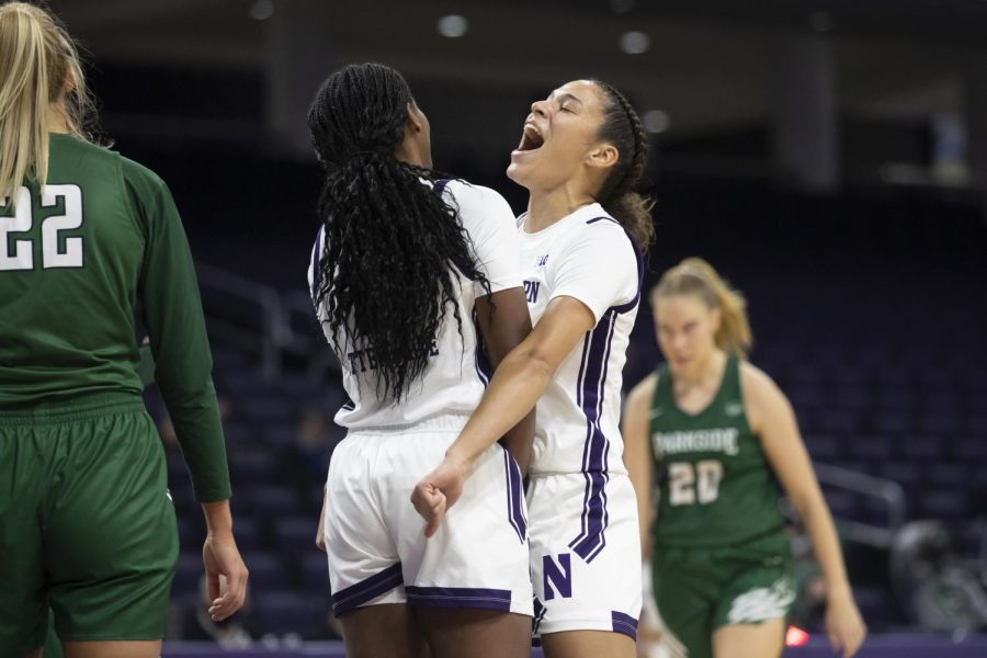 Two+girls+in+white+jerseys+with+purple+stripes+celebrate+on+a+basketball+court.+Two+other+players+in+green+shirts+and+shorts%2C+one+with+the+number+22+and+one+with+the+number+20%2C+are+in+the+background.