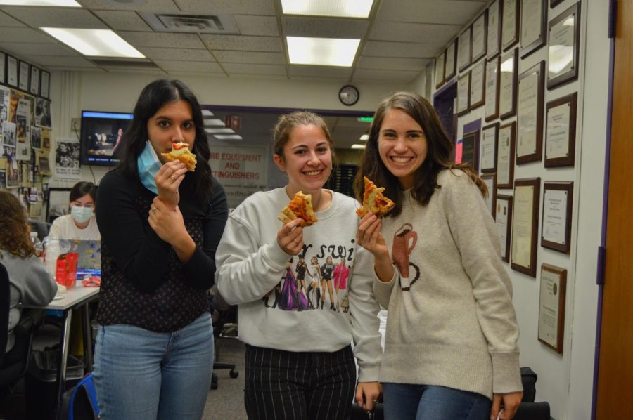 Three people stand together holding pizza in the newsroom.
