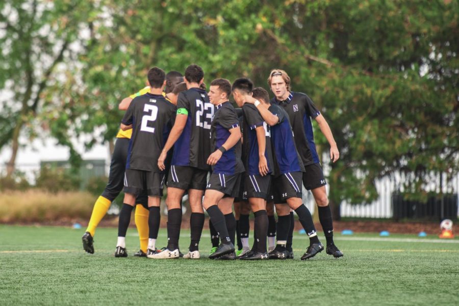 A group of players in black jerseys, black shorts and black socks huddle together on a green soccer field.