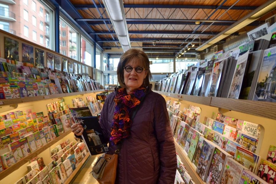 A person with a jacket, scarf and glasses holds two art magazines in the center of an aisle of magazines.