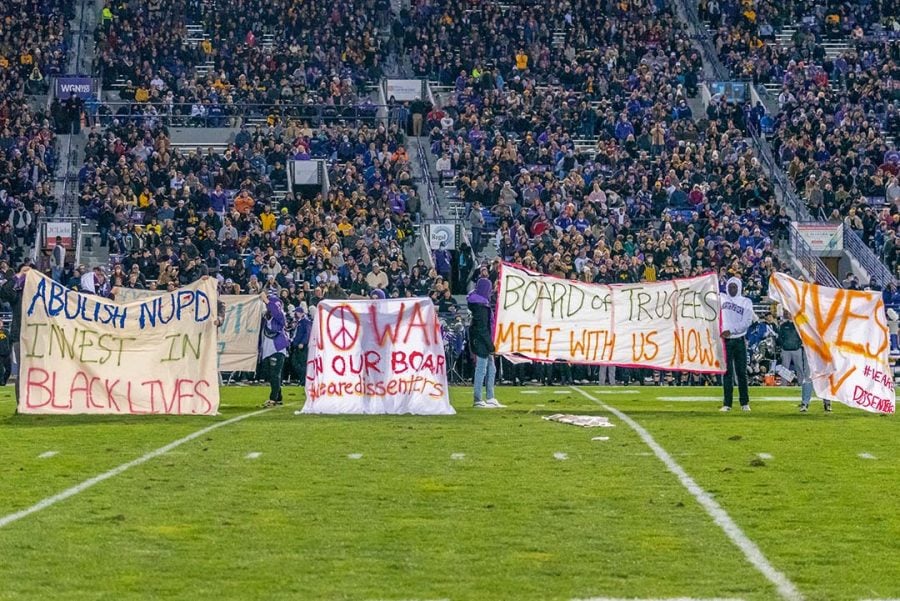 Nine student protesters stand on a football field in front of a stadium of people holding banners that read, “Abolish NUPD invest in Black lives,” “No war on our board #wearedissenters,” “Board of Trustees meet with us now” and “Divest.”