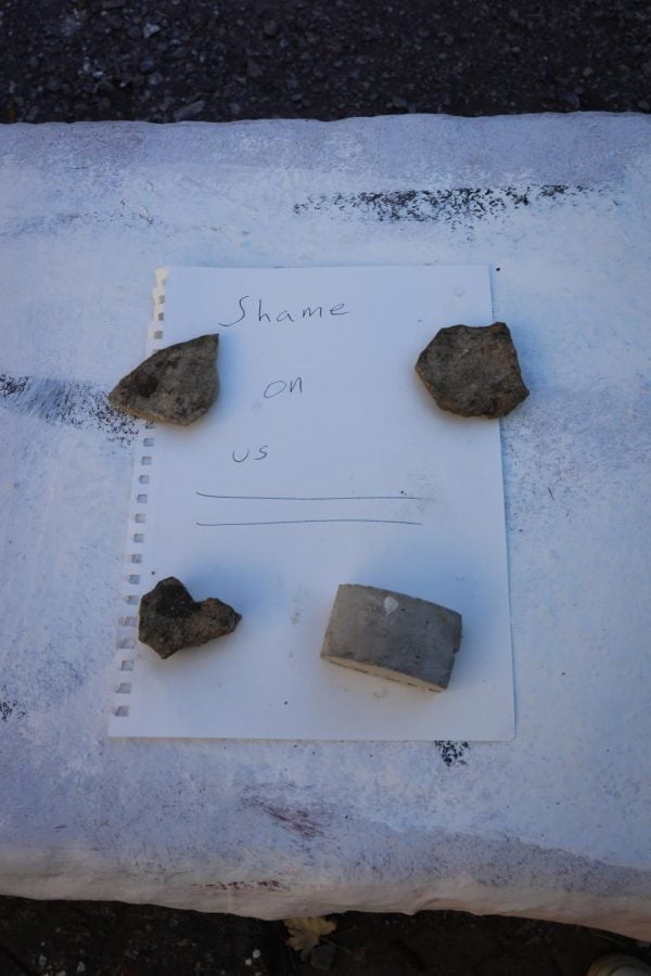 A piece of white paper with “Shame on us” written on it is held down by four rocks.