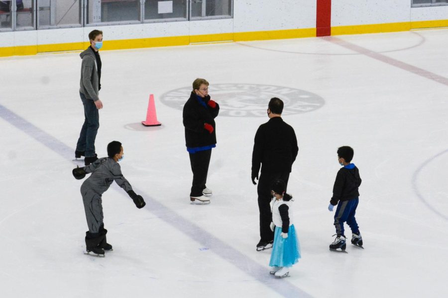 Six people (children and adults) wearing masks skating on an indoor rink.