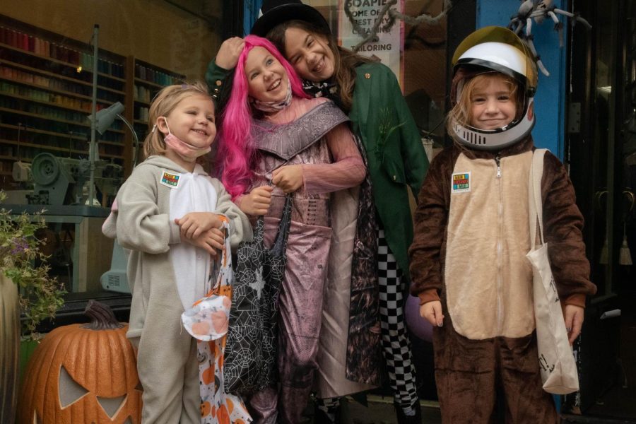 Four kids in different colored Halloween costumes stand in front of a storefront adorned with spooky-themed decorations.