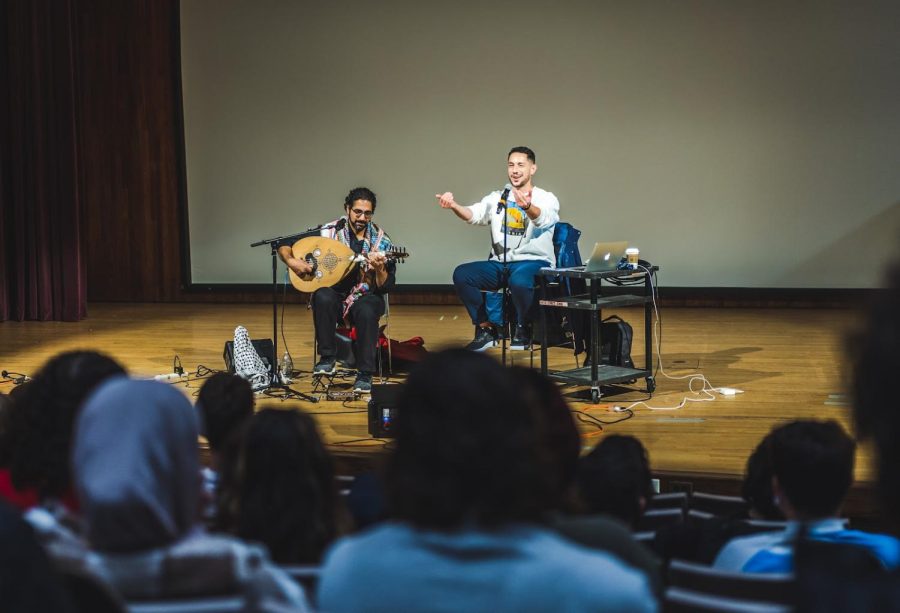 Two individuals on a stage, one singing and one playing the oud, performing in front of a crowd.