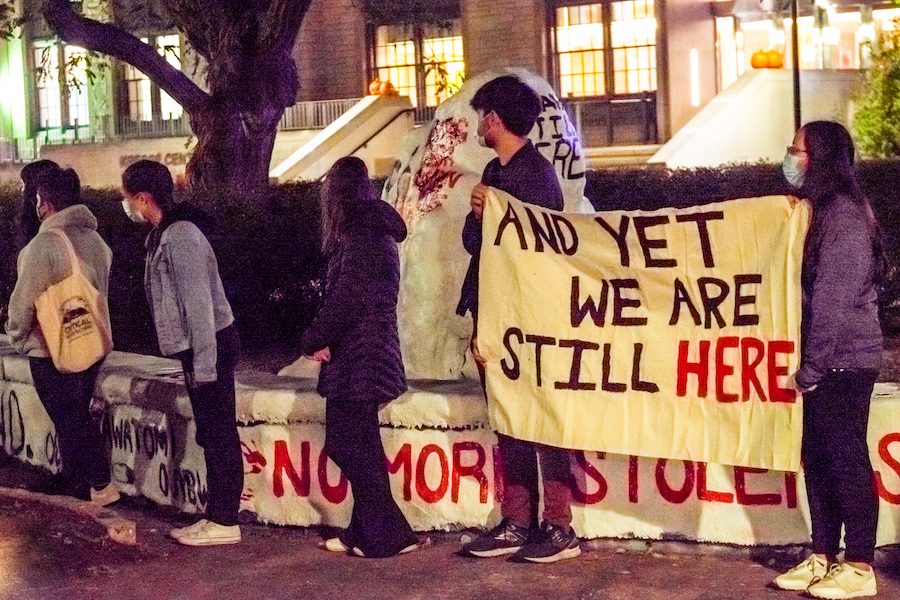 Two students hold a banner reading “And yet we are still here” in front of the Rock.