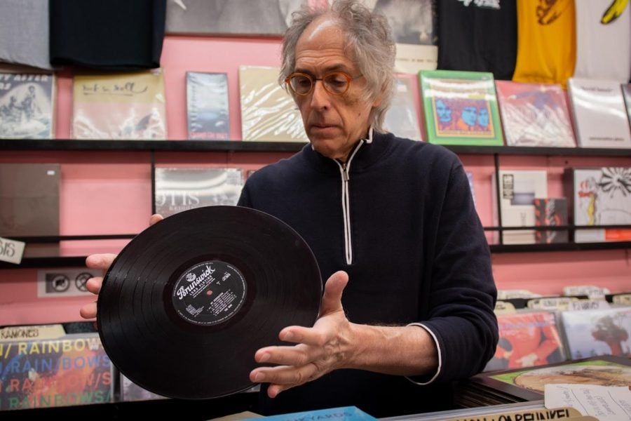 Man in a black sweatshirt holds a black record. Behind him is a pink wall with other colorful titles.