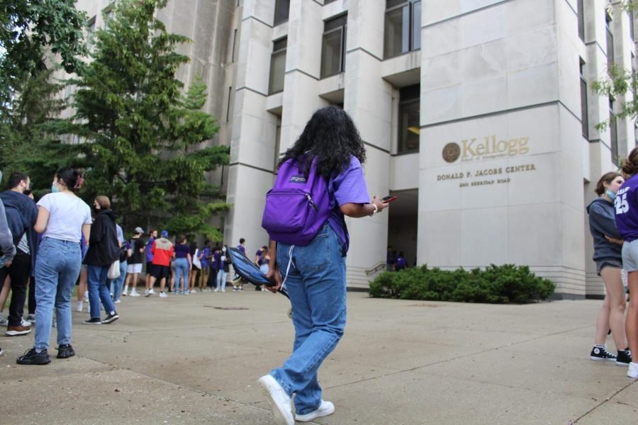 A girl wearing a purple shirt, jeans and a purple backpack walks in front of a large gray stone building.