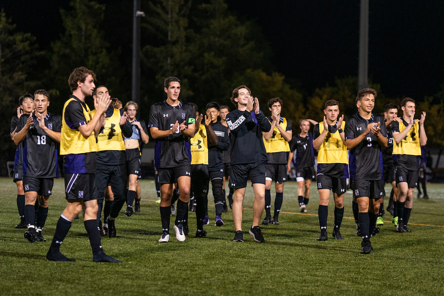 A group of players in black jerseys and black shorts, as well as players in yellow pinnies, black jerseys and black shorts, applaud on a green field.