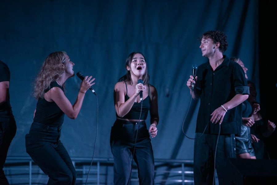 Three people stand on stage, holding microphones with their mouths open.