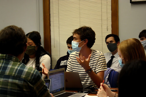 Medill junior Spencer Allan delivers a statement, wearing a blue mask and a black-and-white striped shirt and gesturing with his hands as he speaks. Other Political Union members are seated next to and behind him, also masked.