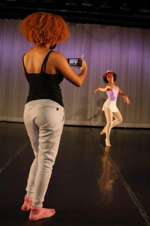 A ballet dancer wearing a crown is photographed by someone holding a cellphone. The two are in a ballroom with warm stage lighting.