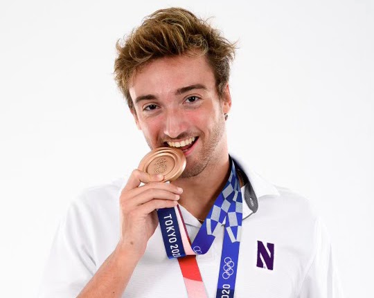 Swimmer in white shirt with two Olympic medals around his neck holds bronze medal in mouth.