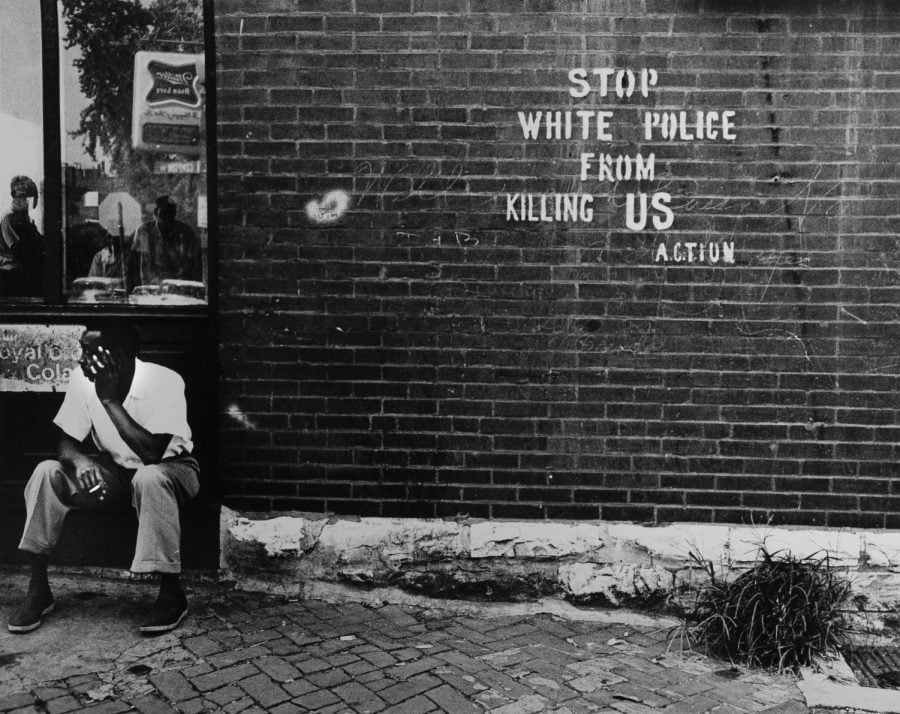 A Black man sits on a bench to the side of a brick wall on which white letters read, “Stop white people from killing us” and “action.” The image is in black and white.