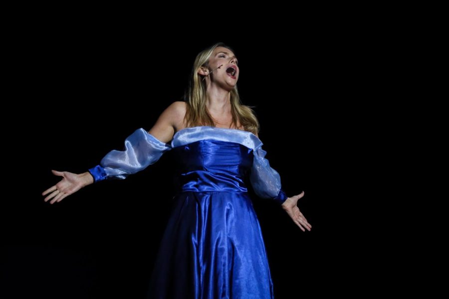 Someone with blonde hair belts on stage in an off-the-shoulder blue gown. A spotlight shines on them. 