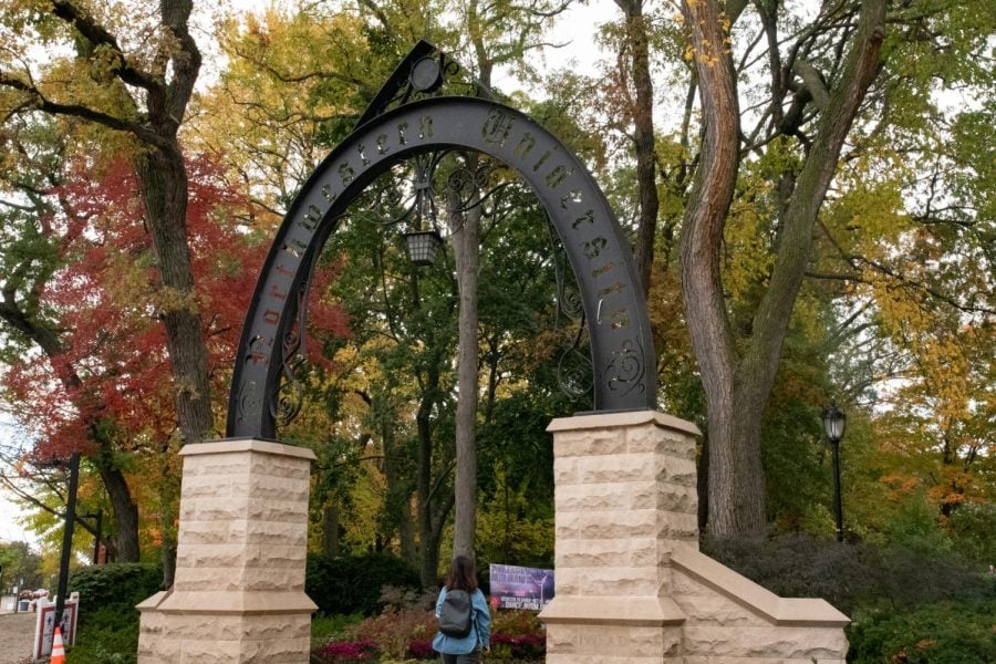 A black arch with the words “Northwestern University” atop of tan stone structures stands among trees with multicolored leaves.