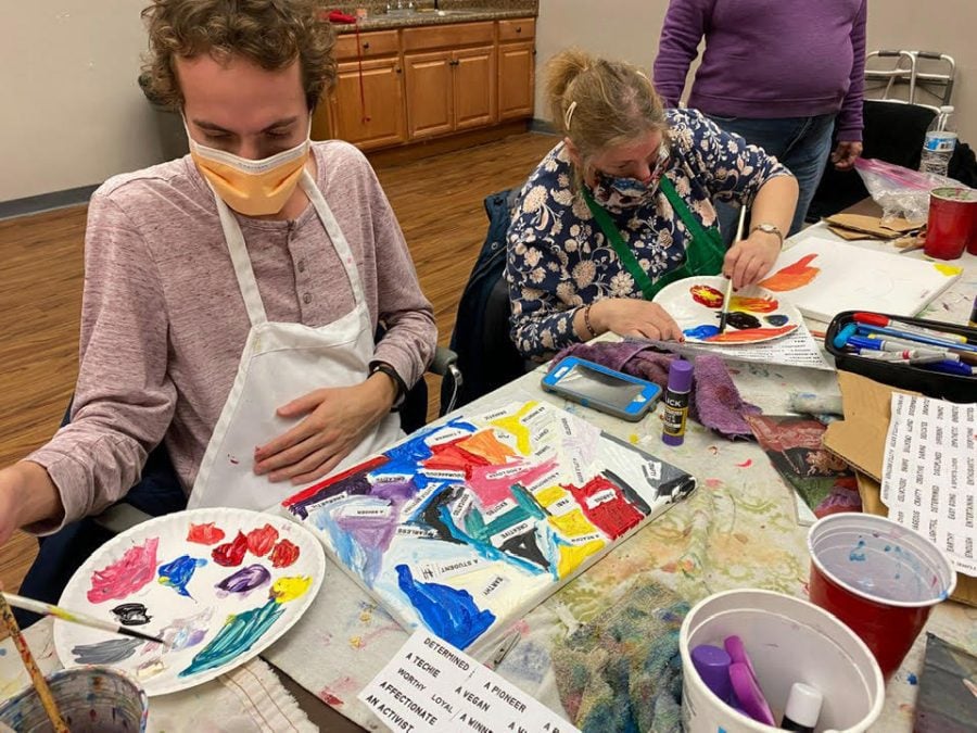 Two Art Club participants sit beside each other at a table covered with art supplies, creating collages out of paint and words printed on paper.