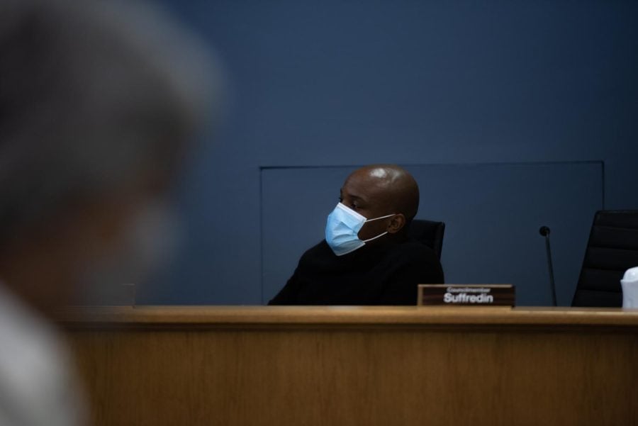 Alderman+Bobby+Burns+sits+at+his+desk+at+City+Council.+He+is+wearing+a+light+blue+disposable+mask+and+black+outfit.+The+background+is+dark+blue.