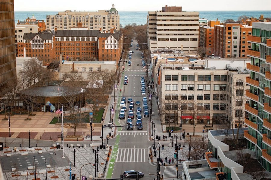An aerial view of Evanston. A wide street runs down the middle with many cars passing through. The street is paved black, the surrounding buildings are brown.