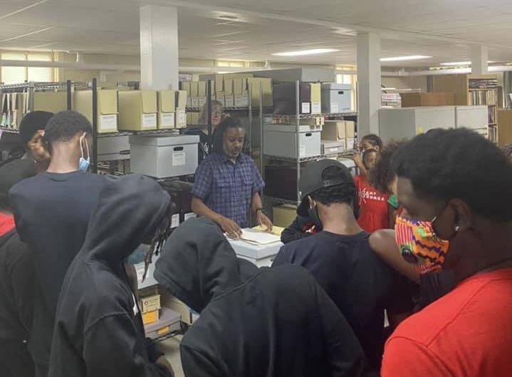 Black students listen to a presentation from a Black man in a room filled with filing cabinets and boxes.