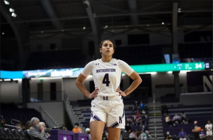 A girl in a white jersey with a purple number 4 on the front stands with her hands on her hips and looks up.