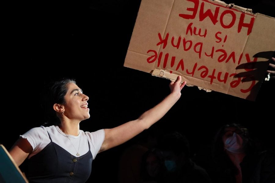 An actress in a white T-shirt and black top smiles as she holds up a cardboard sign upside-down with red lettering.