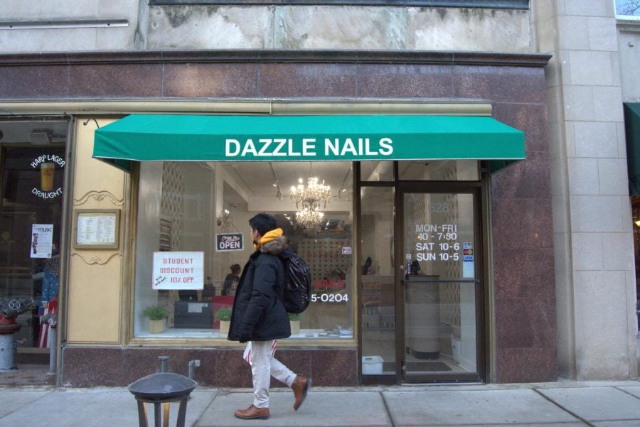 A+person+in+a+winter+coat+walks+past+the+storefront+of+Dazzle+Nails%2C+which+has+a+green+awning+and+glass+windows+which+look+into+an+interior+of+spa+chairs%2C+nail+polish+and+silvery+decor.