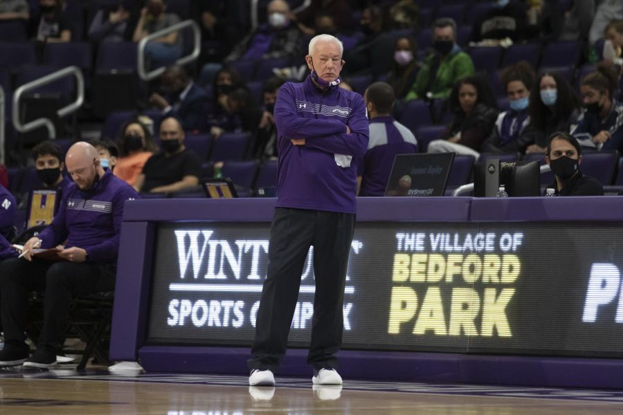Coach in purple shirt and black pants stands on a basketball court with his arms crossed.