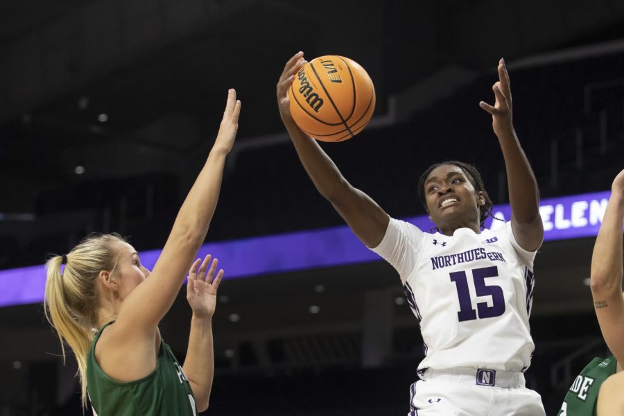 A girl wearing a white jersey with a purple 15 on the front reaches for a basketball in the air while she is facing a girl in a green uniform.