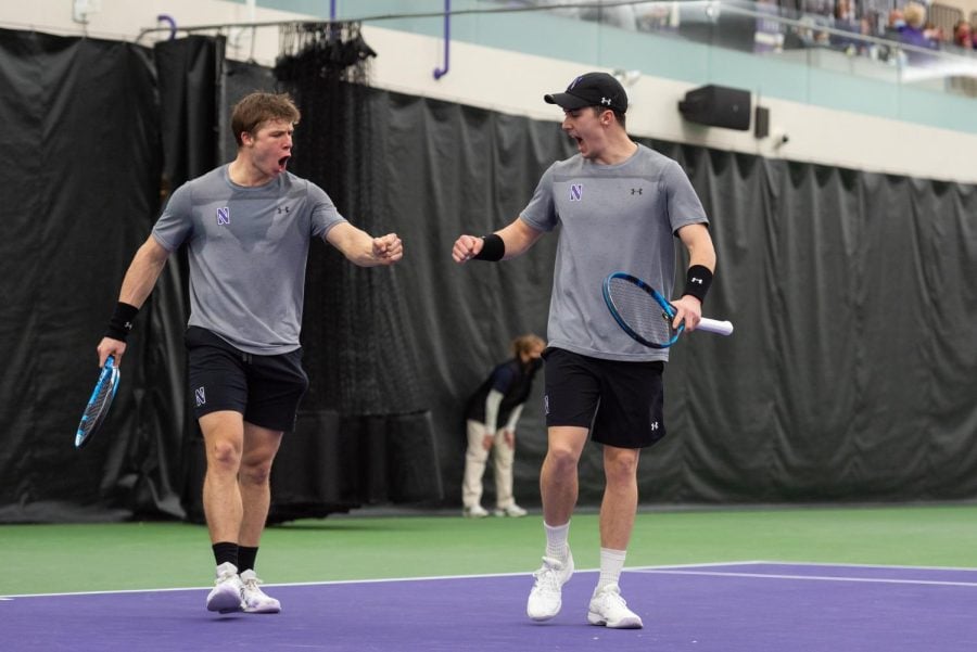 Tennis players in gray shirt and black shorts on purple and green court fist bump with open mouths.