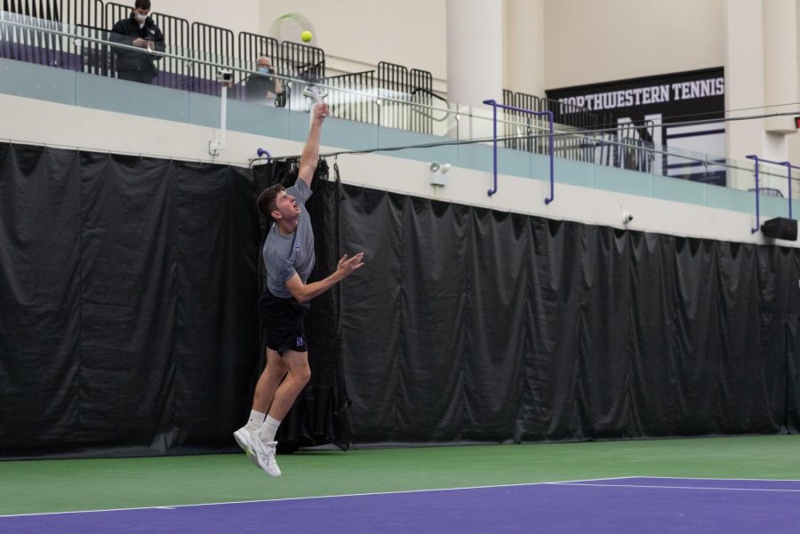 Left-handed tennis player in gray shirt, white shoes and black shorts holds tennis racquet above head to serve on purple and green court.